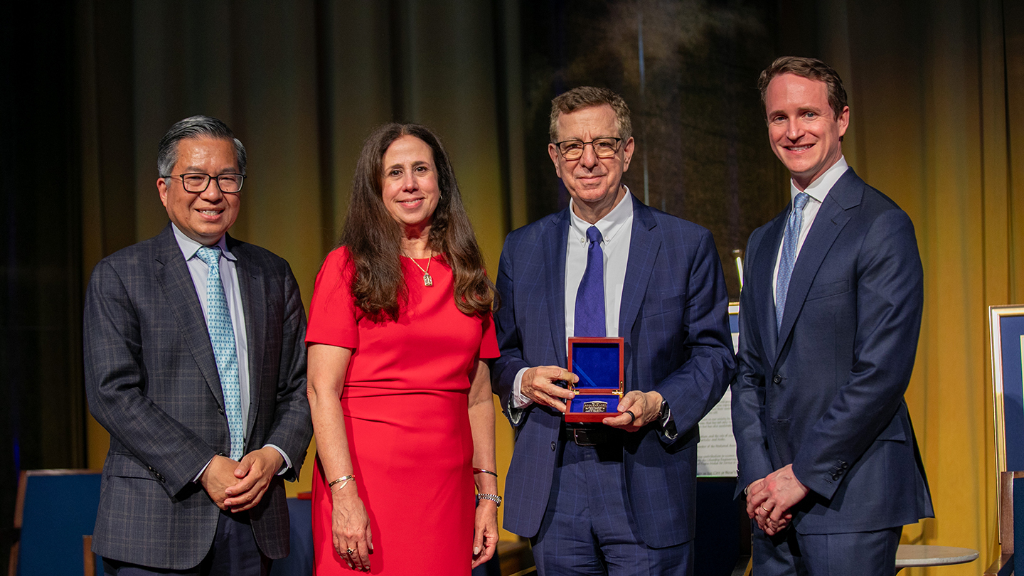 Four people pose onstage at the Columbia Engineering 2026 alumni reunion while one holds an open presentation box with an award medal; framed citations are displayed behind them.