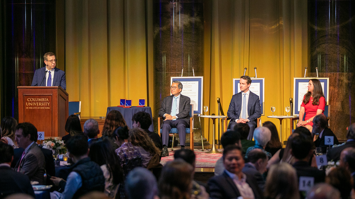 A speaker at a Columbia University lectern addresses a seated audience onstage, with three panelists and framed award displays behind them.