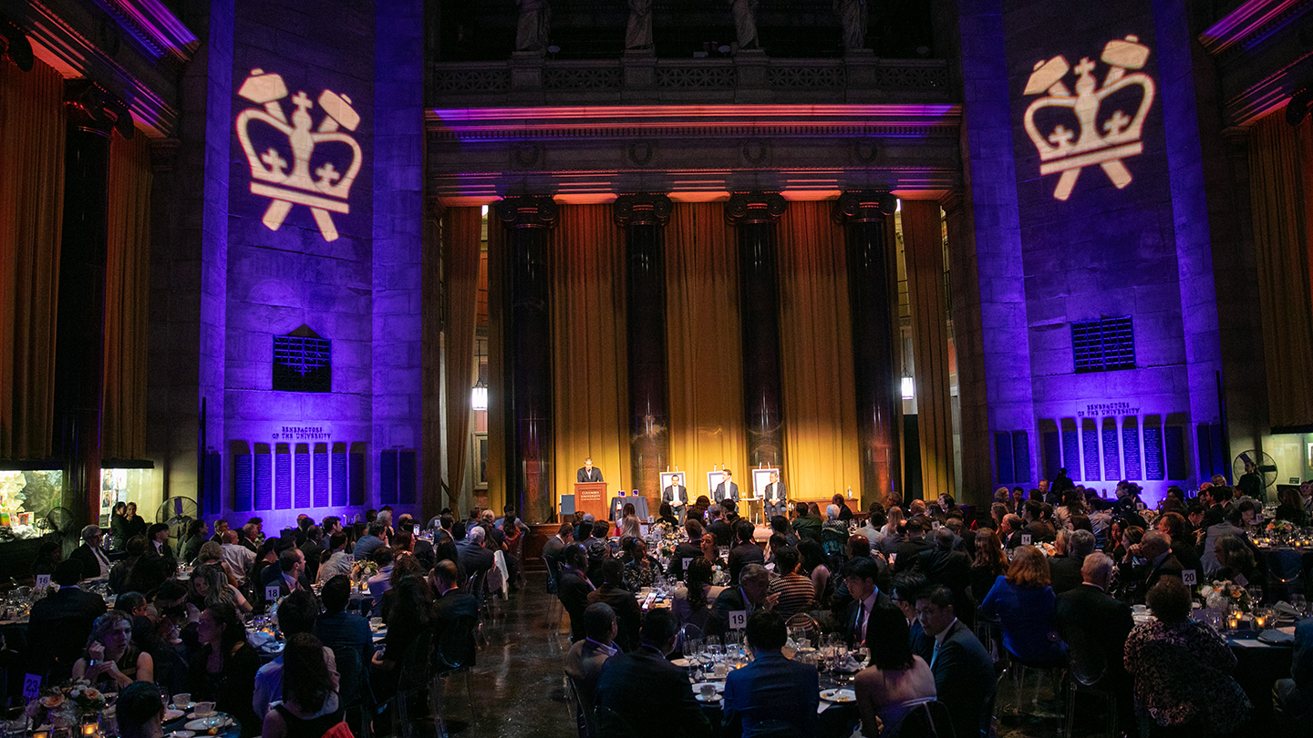 Wide view of a formal dinner in a grand hall lit in purple and gold, with Columbia crown projections on the walls and a stage program at the far end.