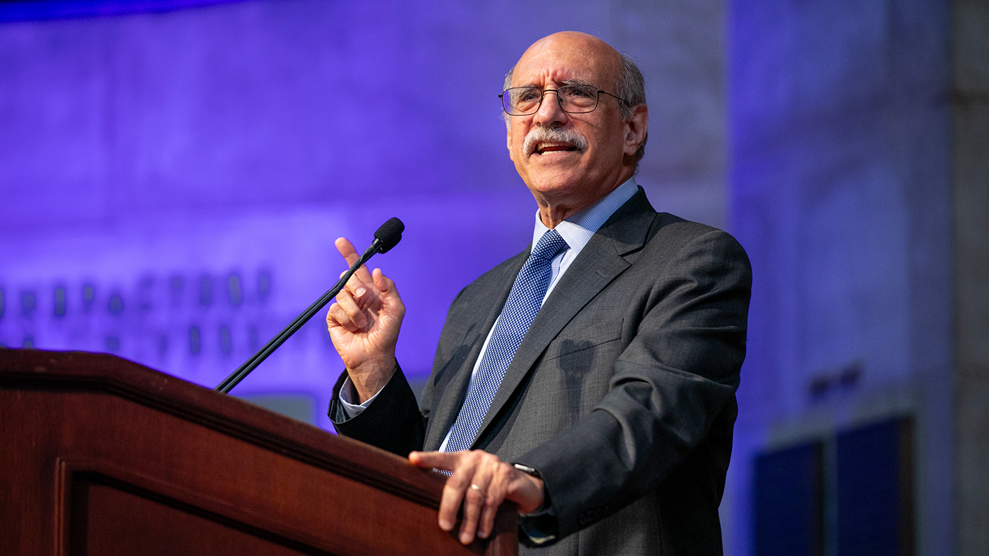 A man in a suit speaks at a podium, gesturing with one hand, against a purple-lit backdrop during the 2026 Columbia Engineering alumni reunion.