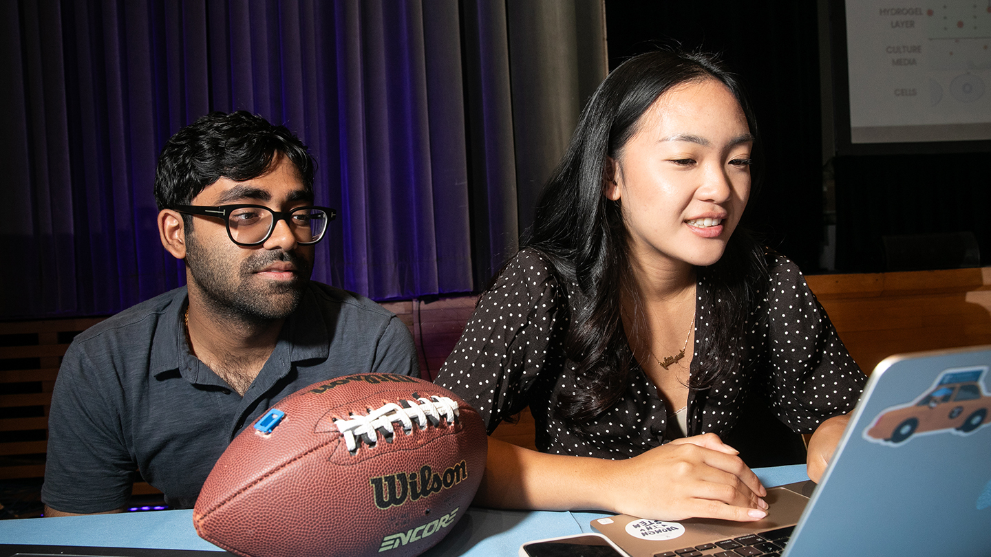 Two students sit at a table looking at a laptop, with a football in the foreground.