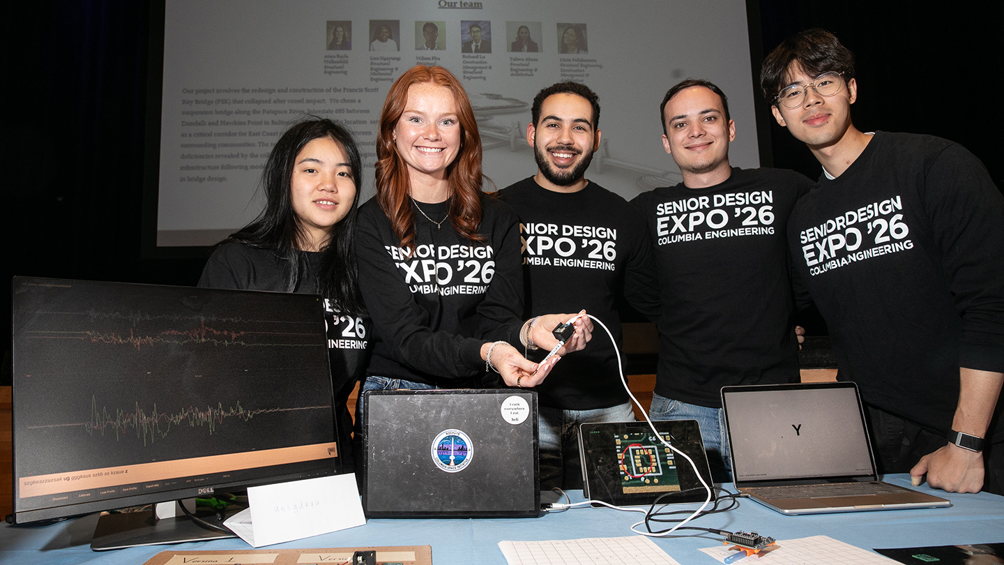 Five students in “Senior Design Expo ’26” shirts pose behind a table of laptops and electronics, with one holding up a sensor device on a cable.sor device on a cable.