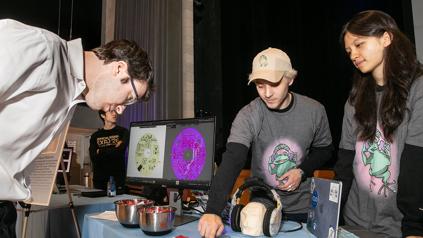 At a demo table, a presenter points to a screen showing circuit-board designs while another student watches and an attendee leans in to look.