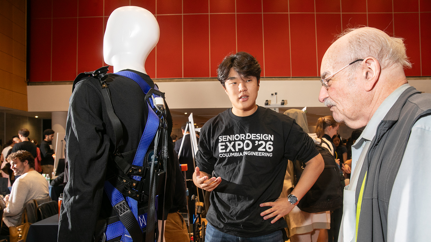 A student in a “Senior Design Expo ’26” shirt explains a safety harness worn on a mannequin to an older attendee.