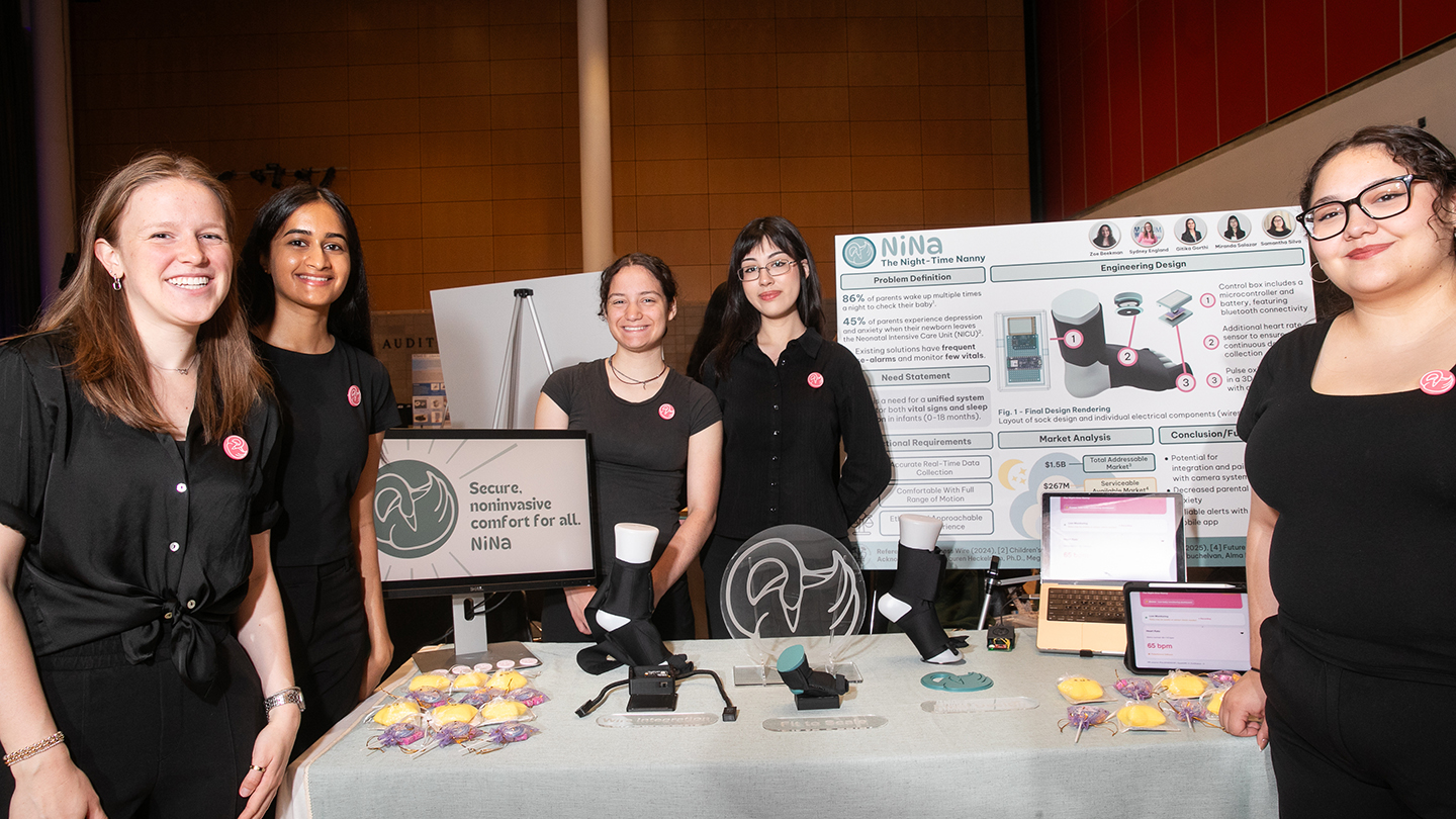 Five students stand behind a table display for “NiNa,” showing prototypes, a laptop, and a monitor with the tagline “Secure, noninvasive comfort for all.”