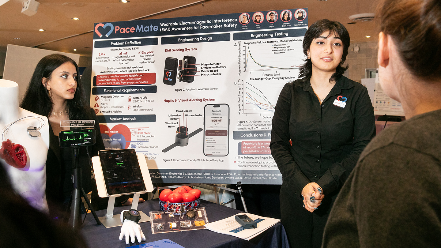 Two presenters stand at a “PaceMate” display about wearable EMI awareness for pacemaker safety, speaking with an attendee across the table.