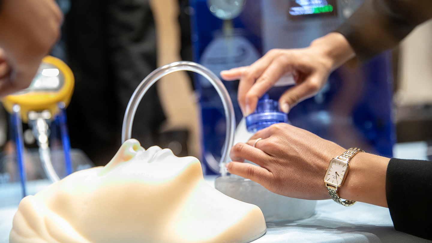 Close-up of hands demonstrating a device with tubing connected to a mannequin head on a tabletop.