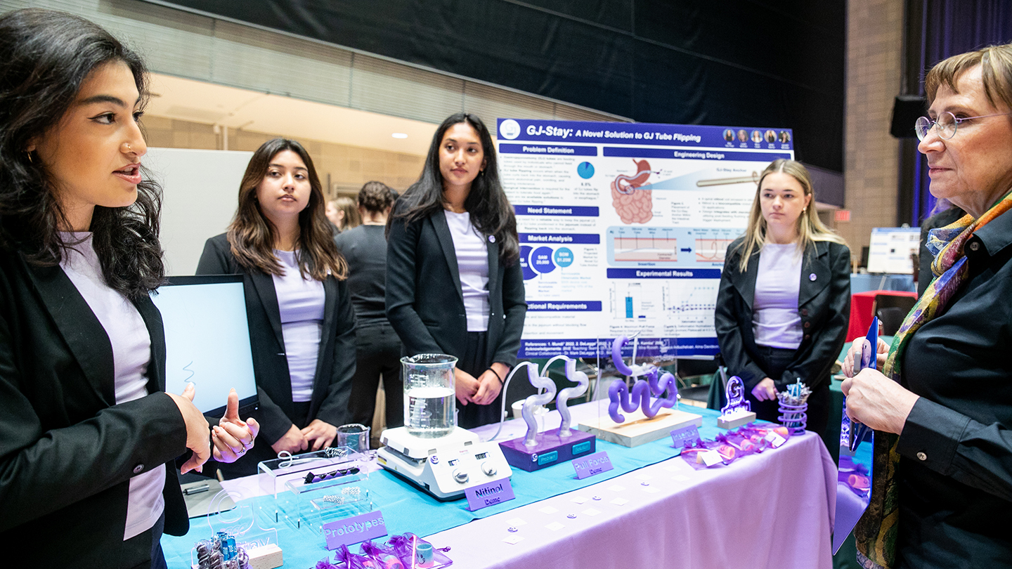 A team presents medical-device prototypes and a project poster to an attendee at a crowded expo table.
