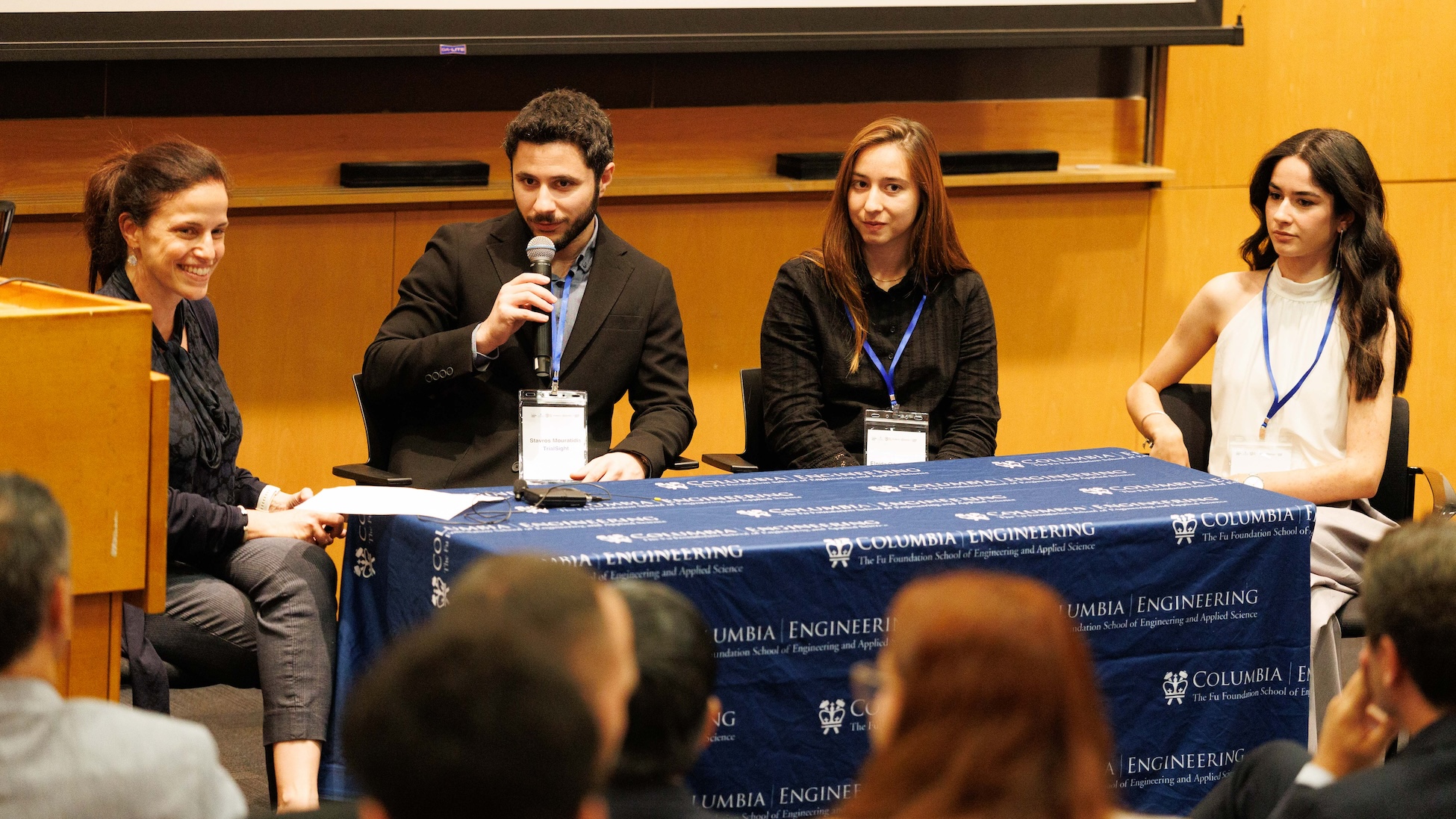 Four panelists sit at a table with a “Columbia Engineering” tablecloth during a talk; one man in a suit speaks into a handheld microphone while three women listen, with an audience visible in the foreground.