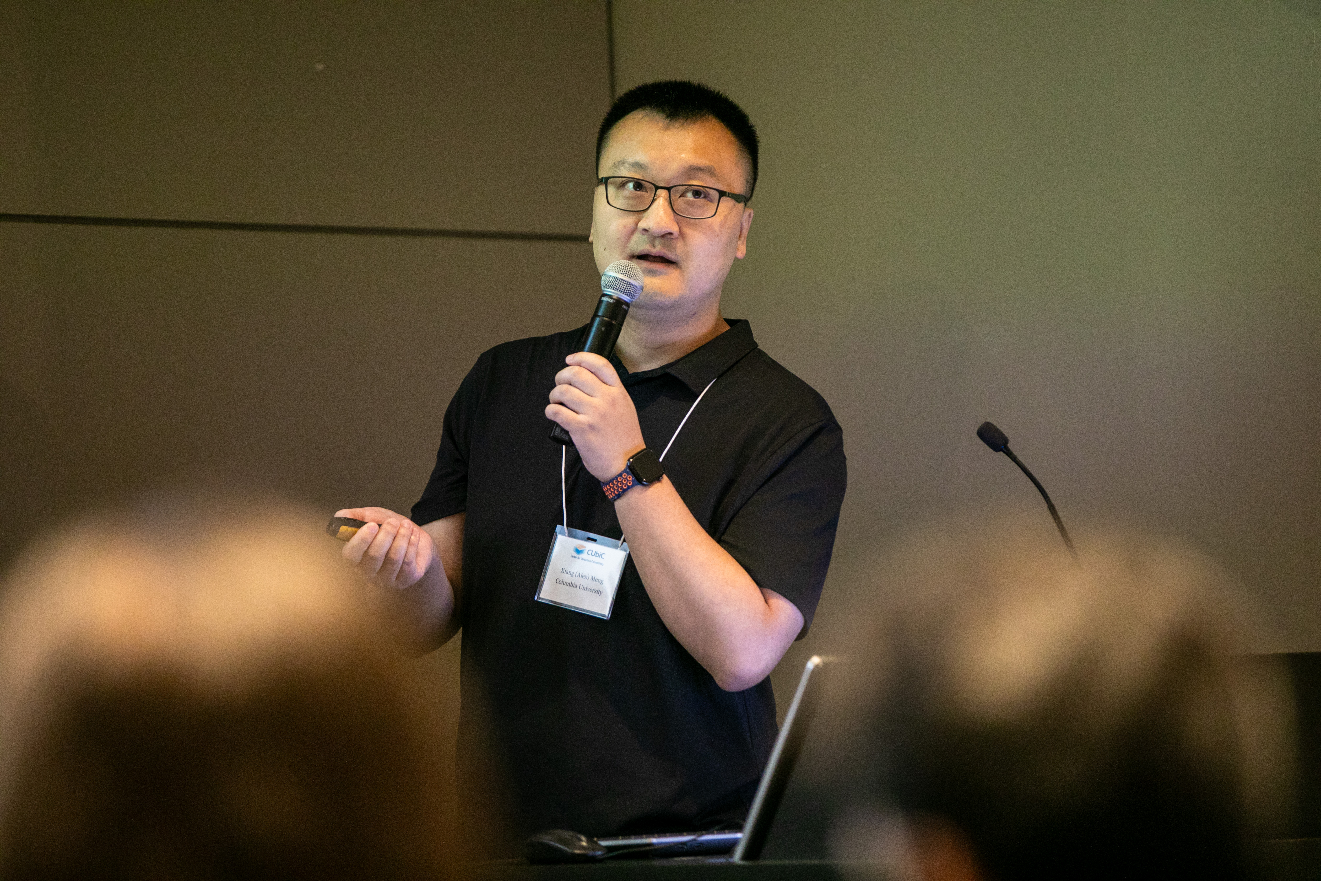 A conference presenter in a black shirt holds a microphone and remote, speaking to an audience.