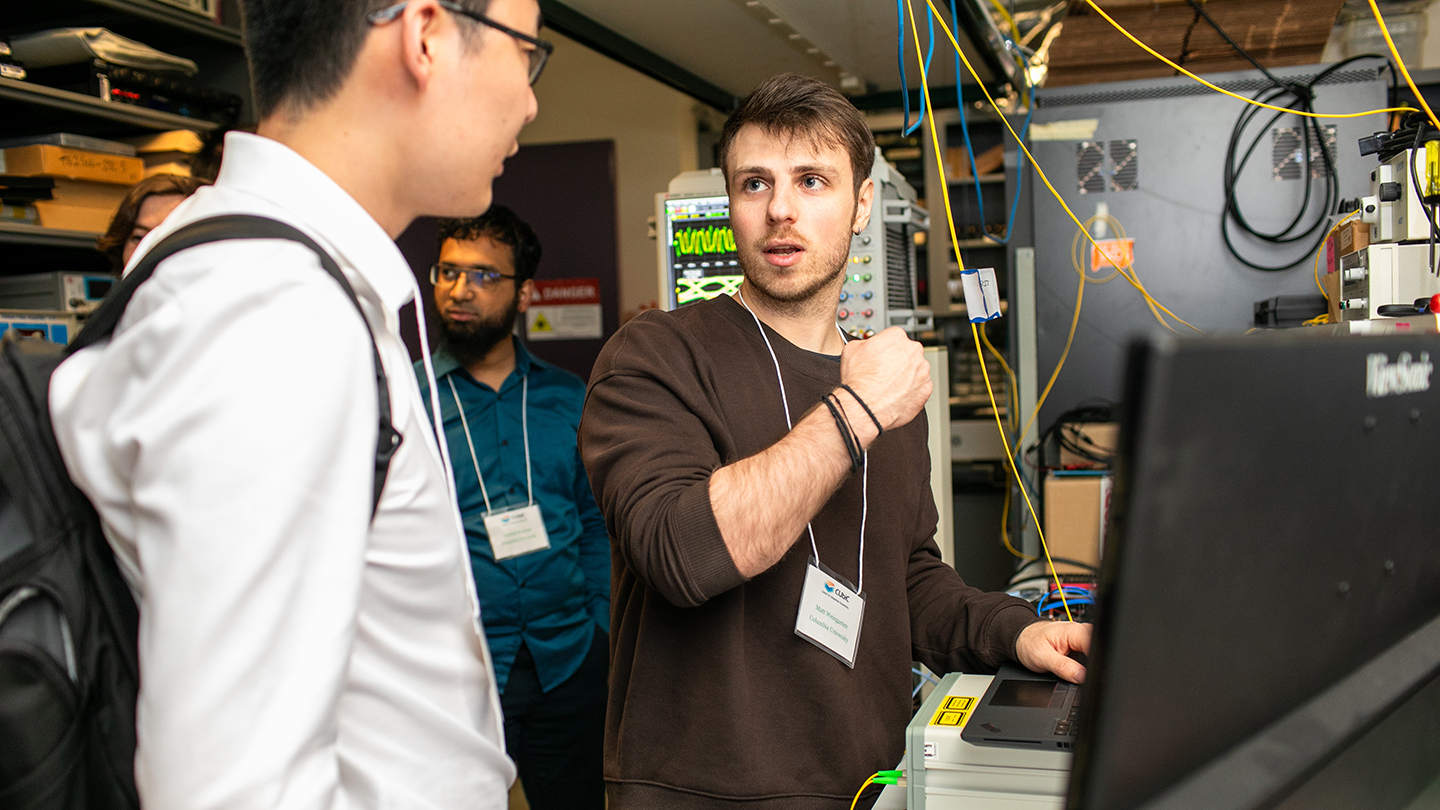 In a lab filled with equipment and cables, Matt Weingarten explains something at a workstation to two visitors.