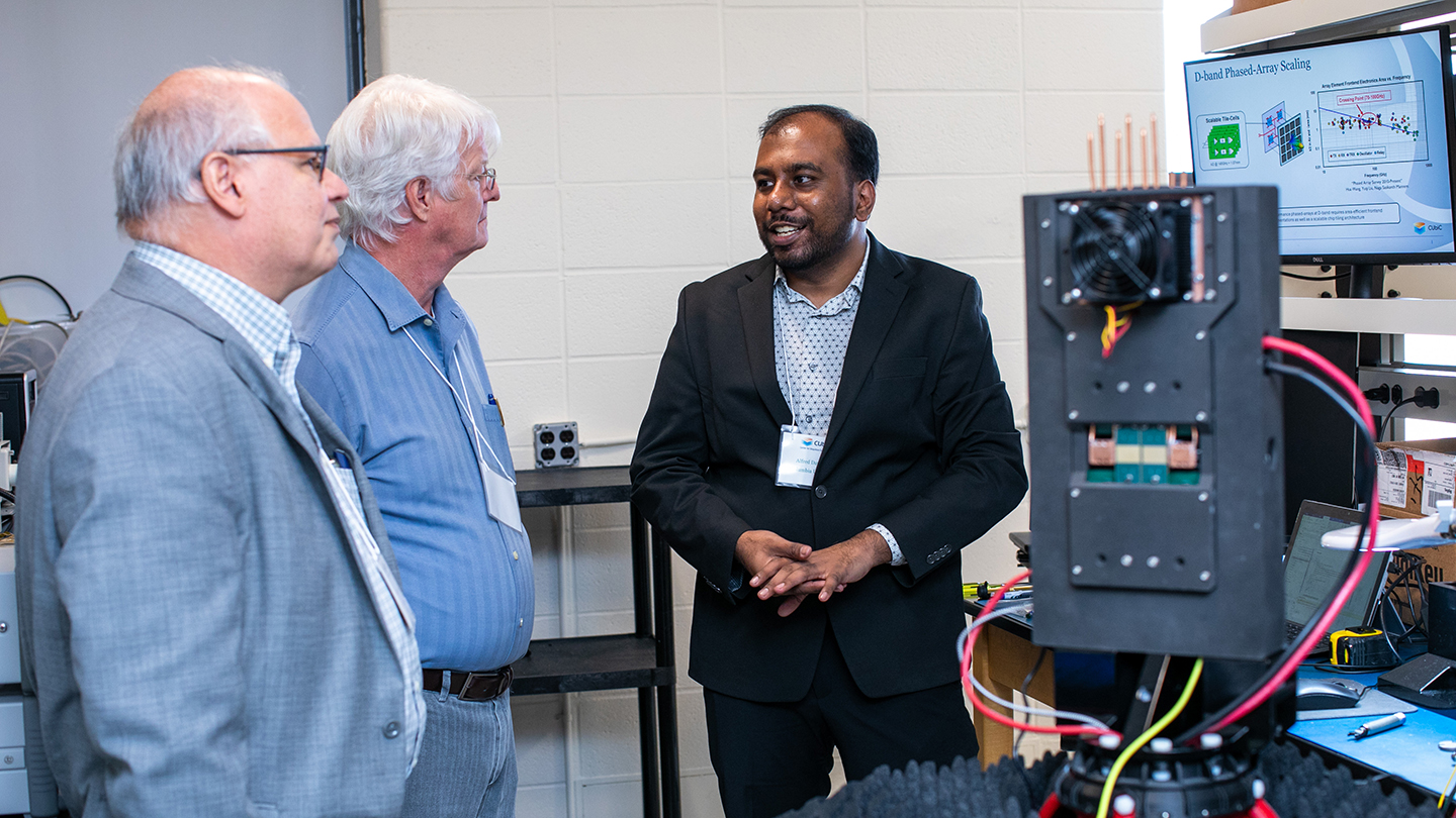  In a research lab, three conference attendees talk beside a piece of electronic test equipment while a monitor displays a CUbiC technical slide.