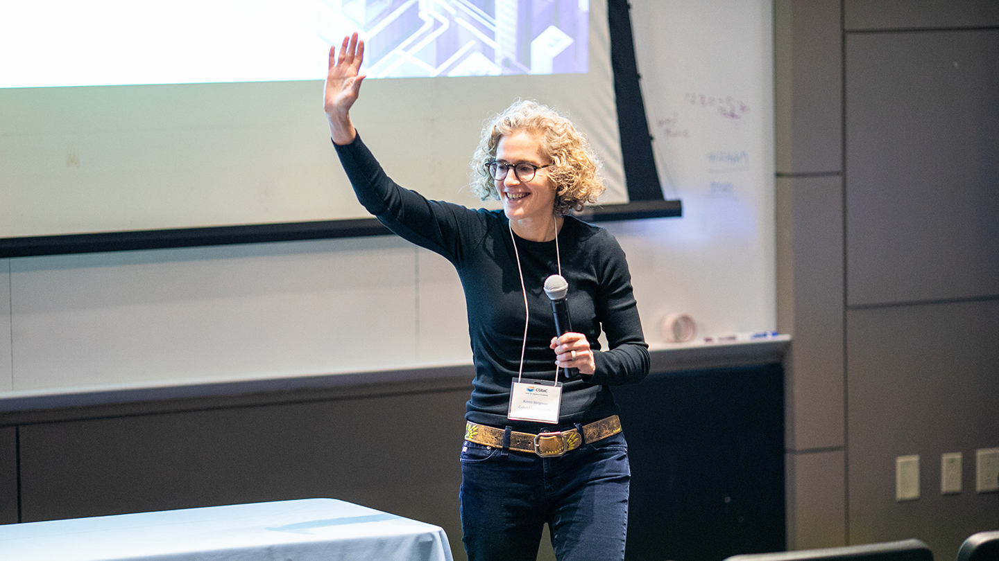 Keren Bergman holds a microphone and waves to the audience in front of a projected slide.