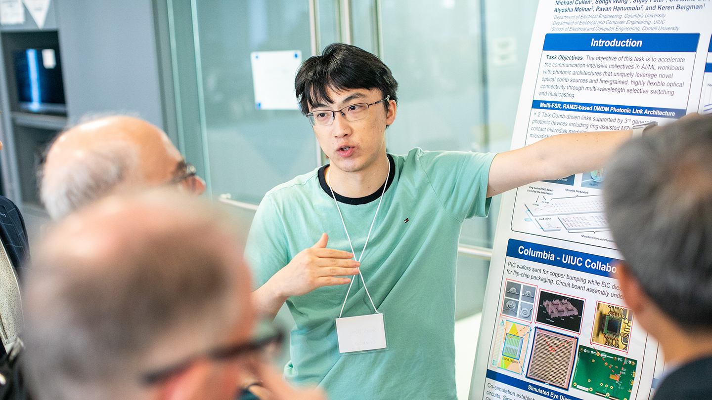 A student presenter gestures toward a research poster while explaining it to attendees gathered nearby.
