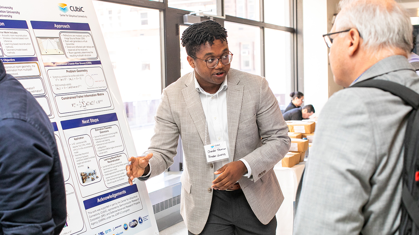 A researcher stands beside a CUbiC poster, talking with two attendees in a bright atrium-like space.