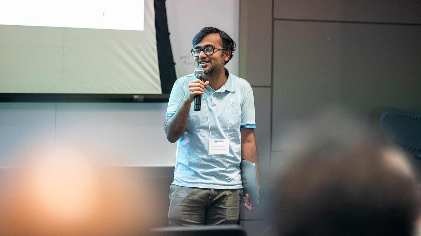 A presenter in a light polo shirt smiles while holding a microphone in a lecture room.