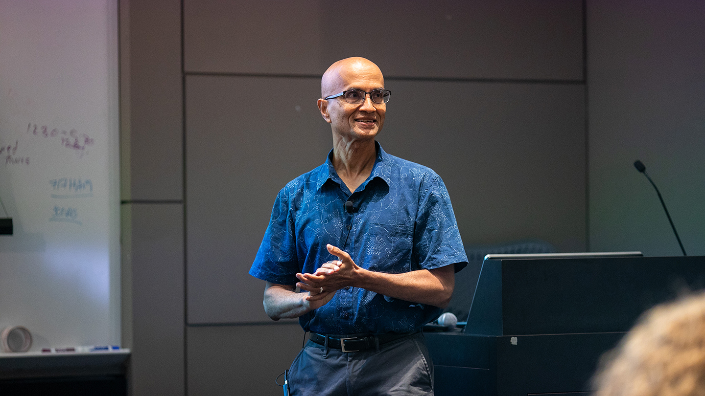  Naresh Shanbhag stands near a lectern, hands clasped, as if pausing between points during a talk.