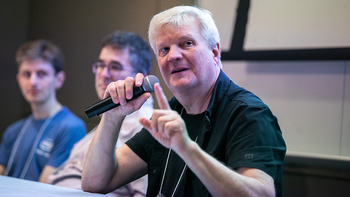  Zoran Zvonar holds a microphone and raises a finger while making a point; other panelists sit beside him.