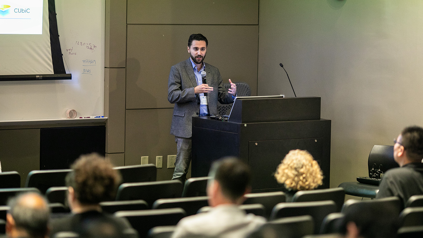 Wide shot of a Brett George at a podium speaking to an audience seated in a lecture hall; a CUbiC slide is projected.
