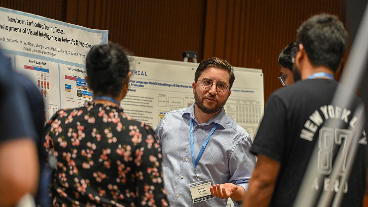 A poster presenter wearing a name badge explains a research poster to several attendees gathered closely in conversation.