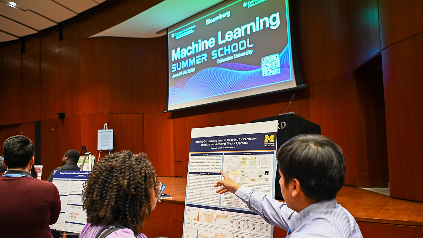 At Columbia University’s Machine Learning Summer School, two attendees review a research poster; one points at figures on the poster while a large “Machine Learning Summer School” slide is projected above the auditorium stage.