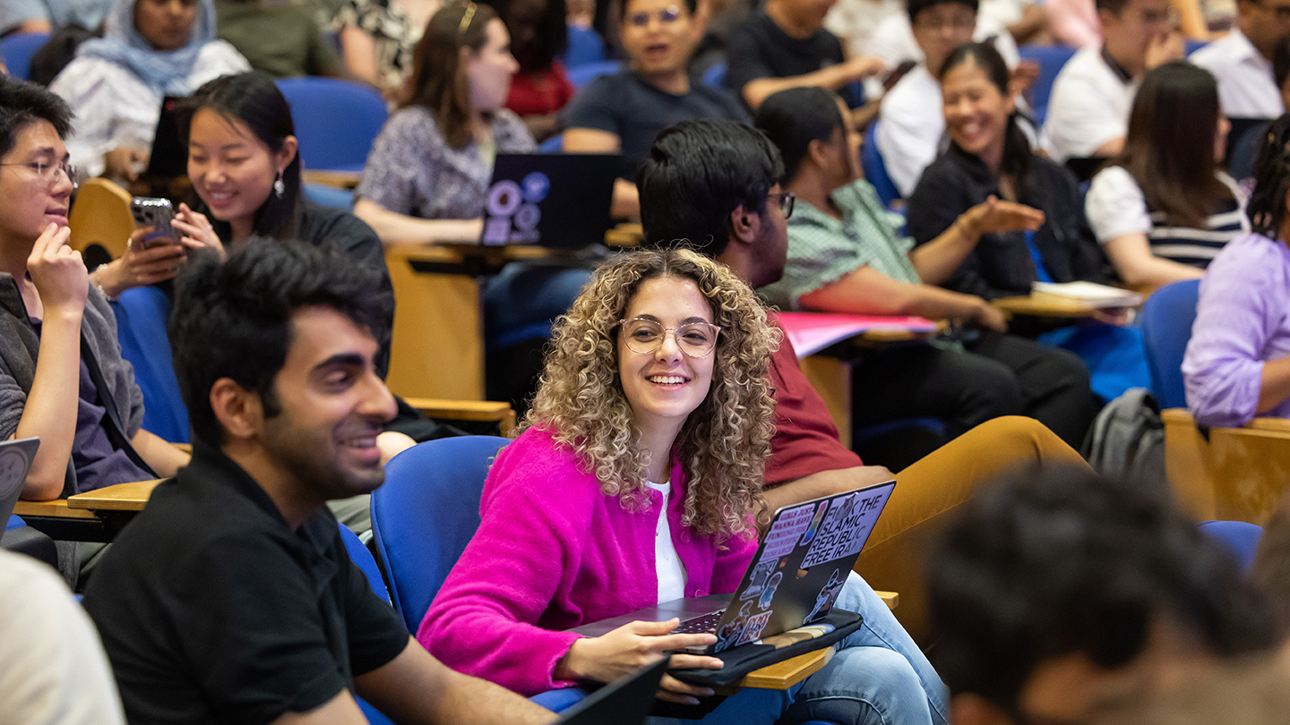  Audience members seated in a lecture hall at Columbia’s Machine Learning Summer School chat and smile before a session; one attendee in a bright pink cardigan looks toward the camera while holding an open, sticker-covered laptop.