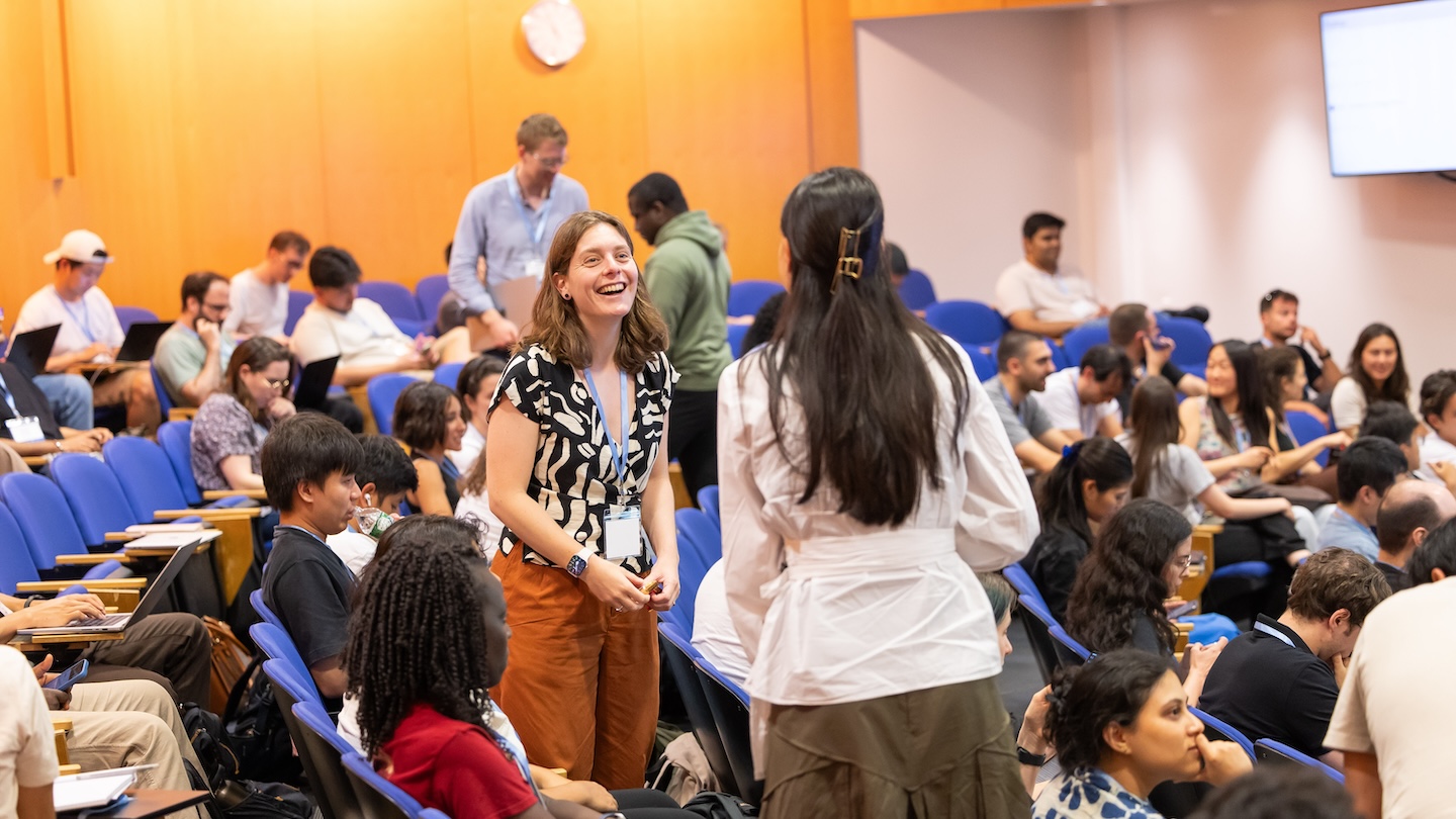 In a lecture hall at Columbia University’s Machine Learning Summer School, attendees sit in blue seats with laptops and notebooks while two participants stand in the aisle chatting; one smiles and laughs as others settle in around them.