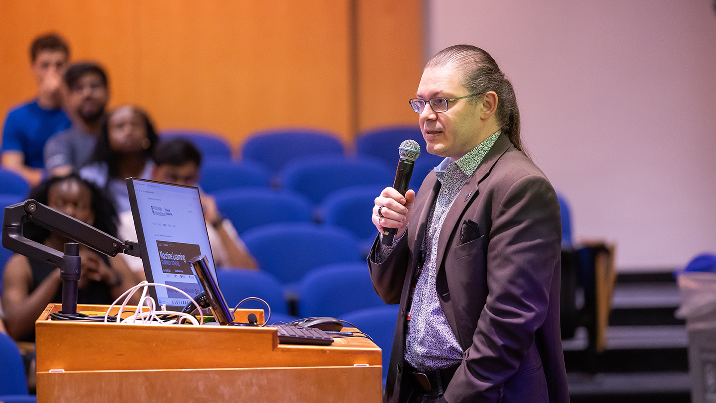 Gary Kazantsev speaks into a handheld microphone beside a laptop at a lecture podium, with audience seating blurred in the background.