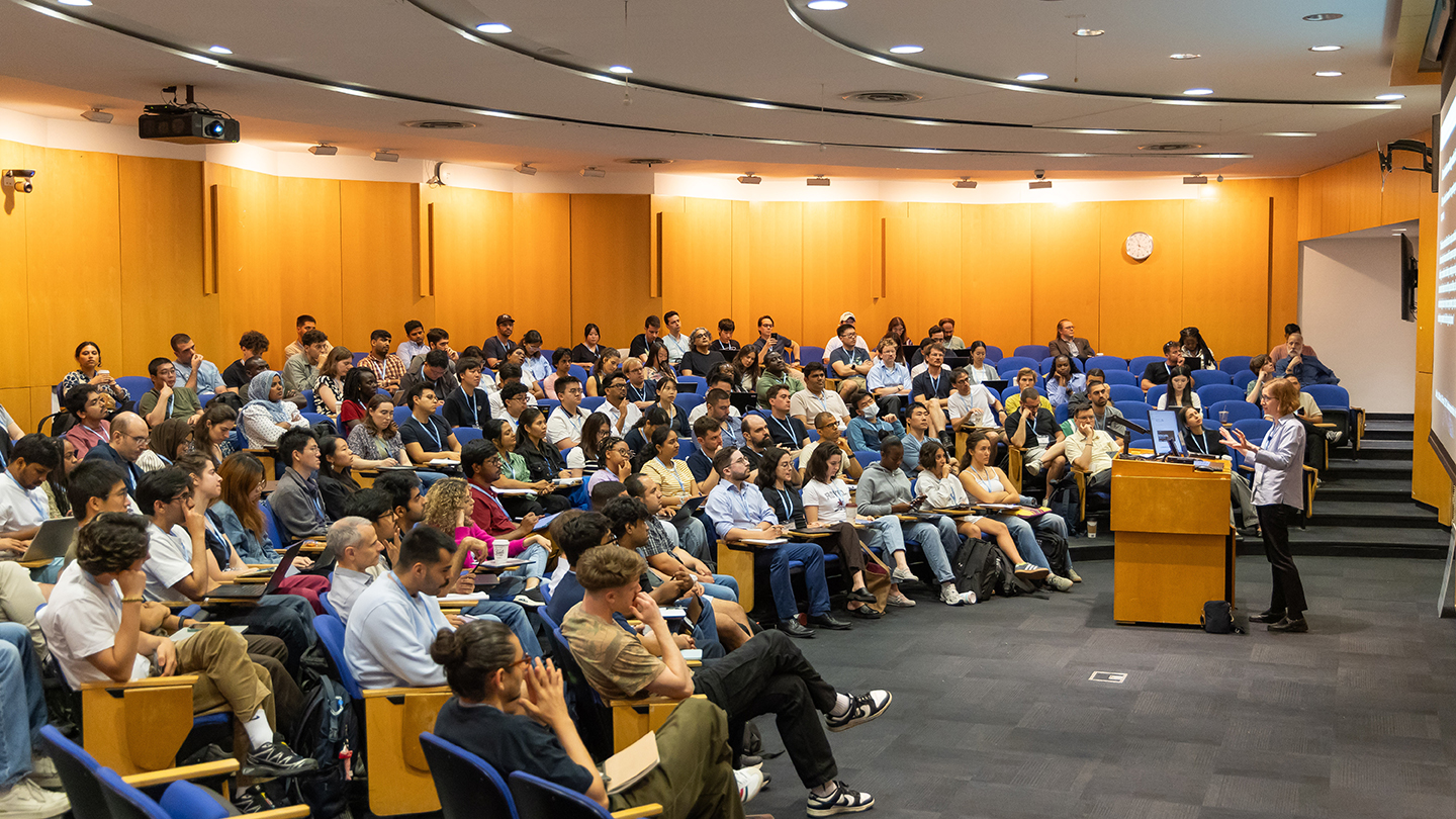Wide view of a packed auditorium during a talk at Columbia’s Machine Learning Summer School, with participants seated in rows facing the presenter and projection screen.