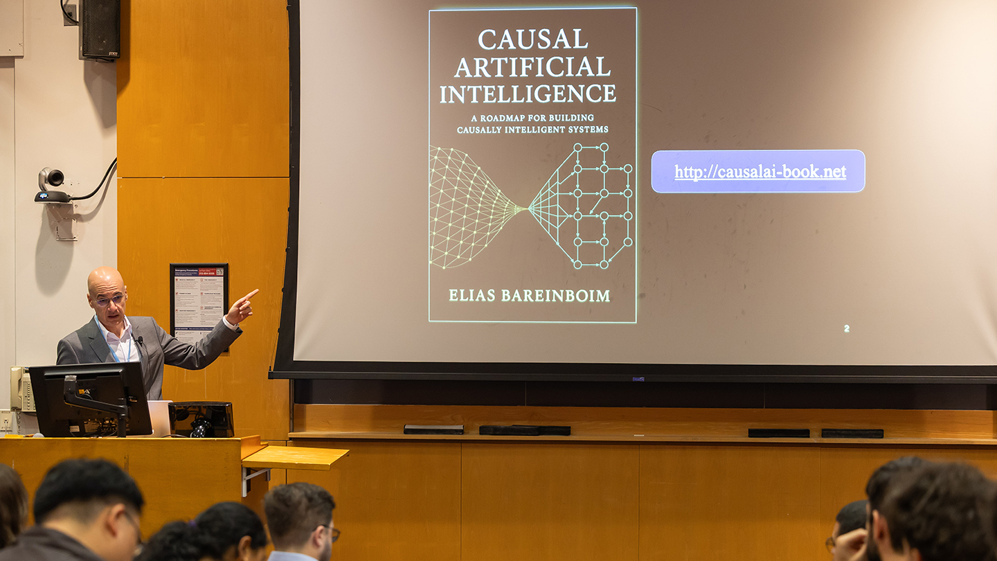 Elias Bareinboim gestures toward a projected slide featuring the book cover “Causal Artificial Intelligence” and a web link, while the audience looks on.