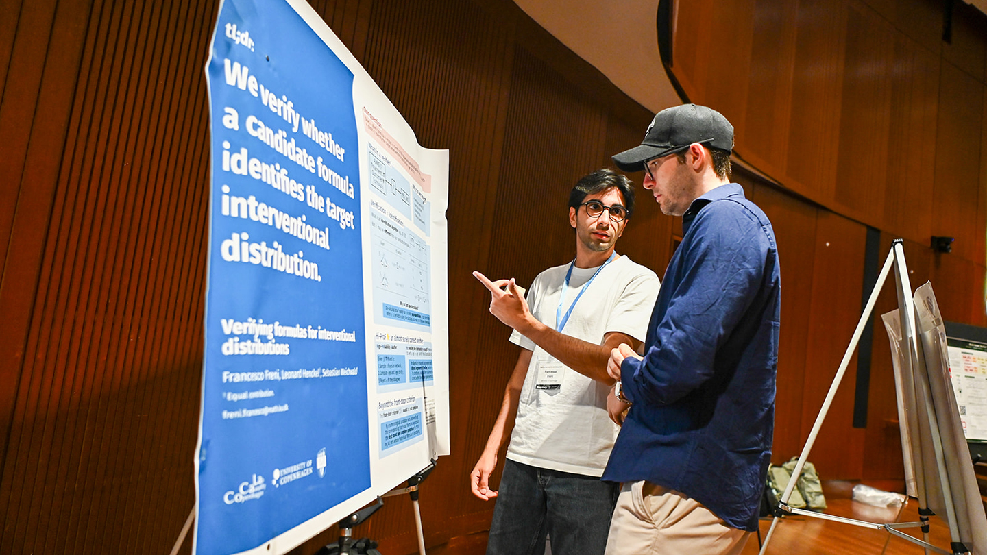 Two attendees wearing lanyards discuss a research poster on an easel; one points at the poster while the other listens.