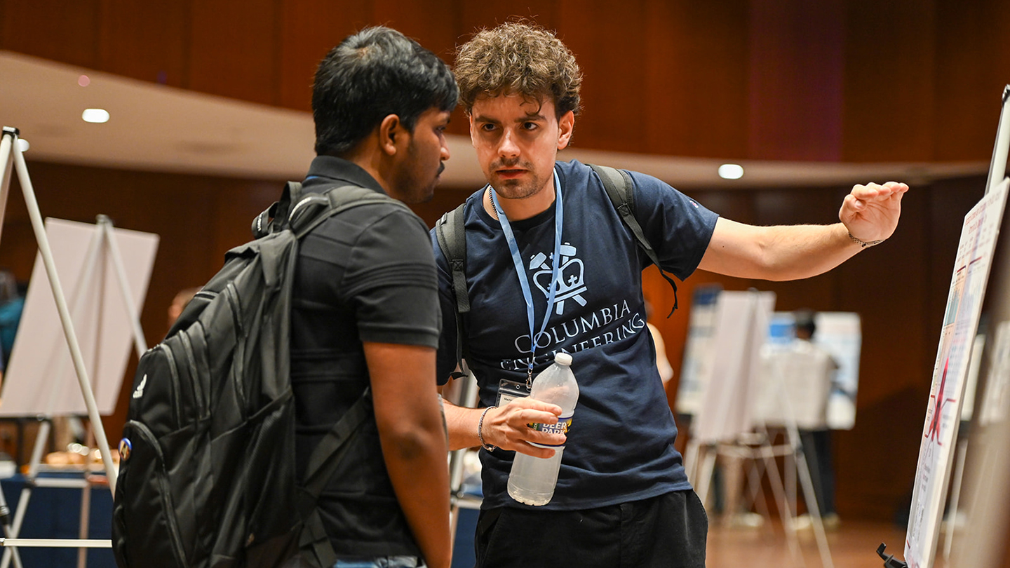  At the poster session, an attendee with a backpack holds a water bottle and gestures toward a poster while speaking with another attendee.