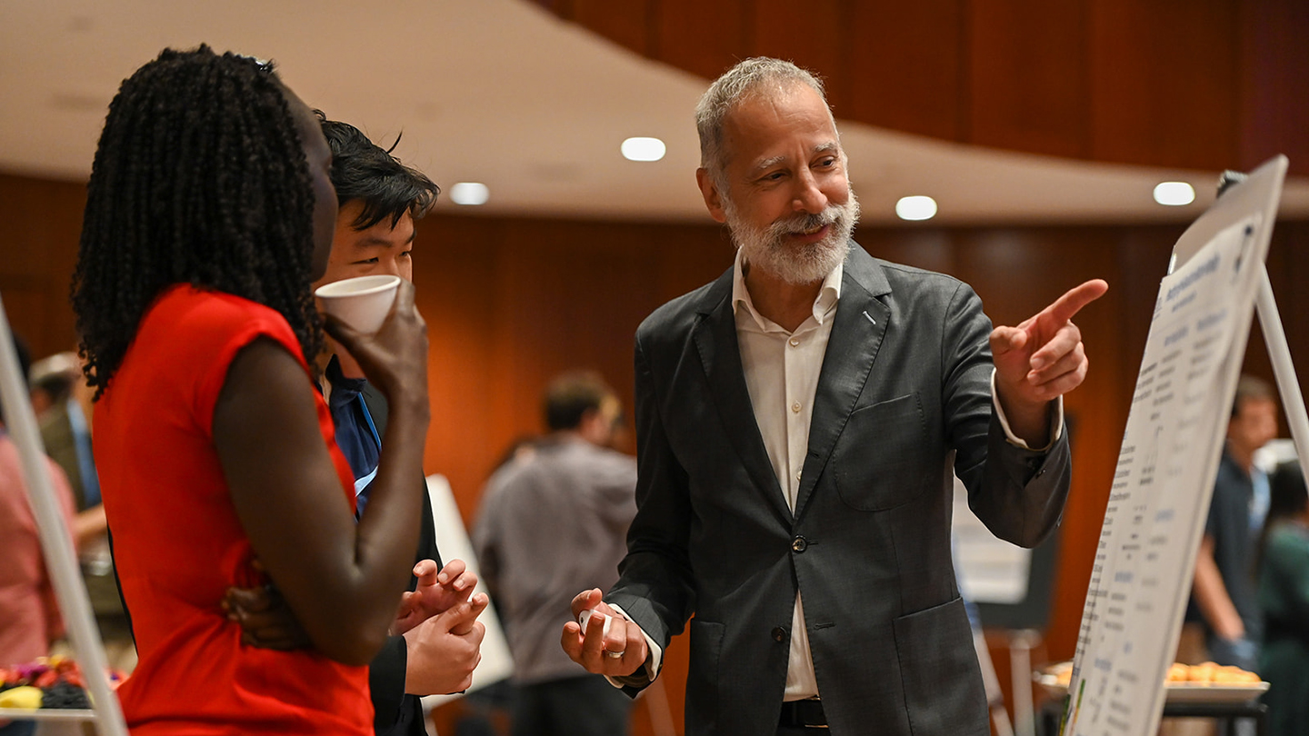 Ali Hirsa points at a poster while talking with two attendees, one holding a cup, in a busy poster-session space.