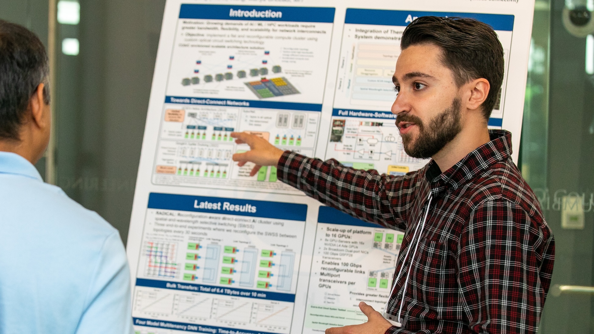 A man in a red-and-black plaid shirt presents a research poster, gesturing toward charts and diagrams while speaking to another person in the foreground at what appears to be a conference or poster session.