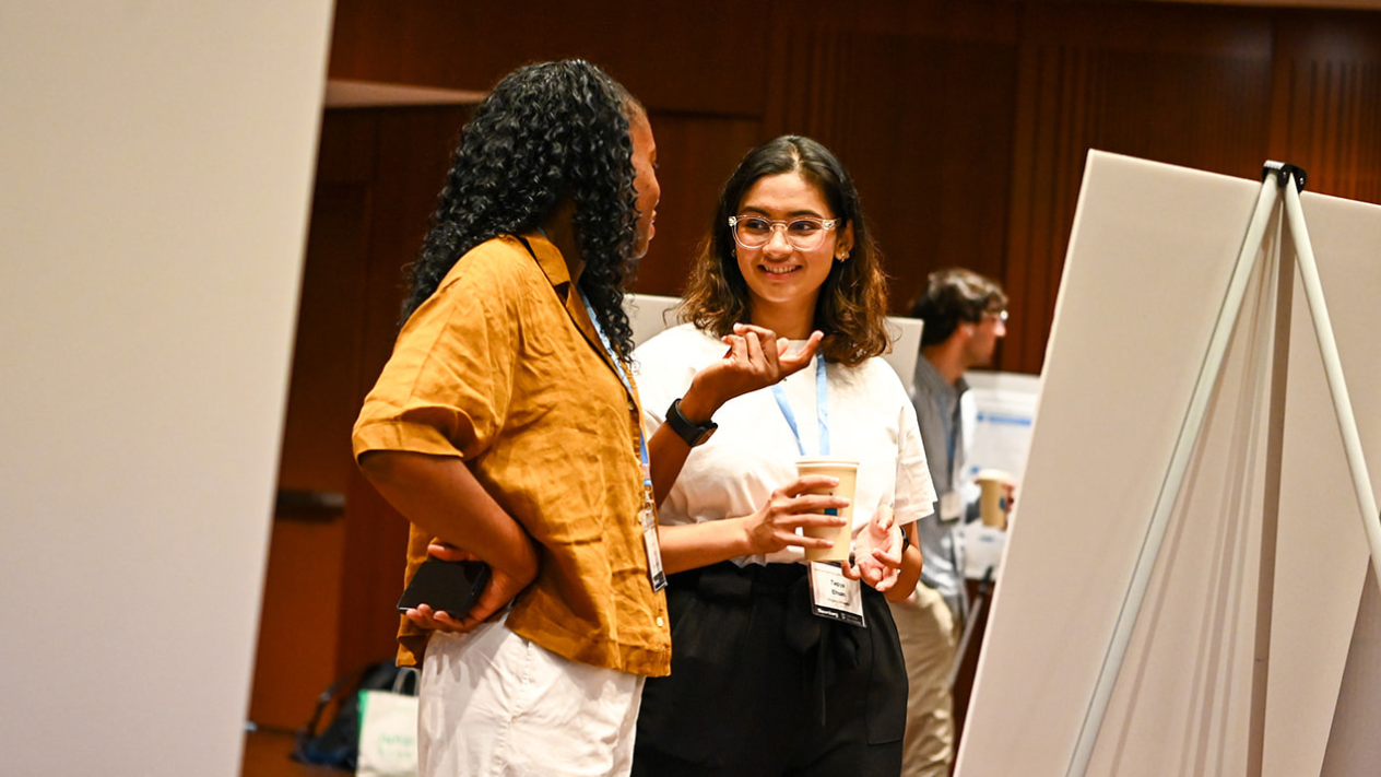Two conference attendees wearing lanyards stand by a white poster board on an easel, talking and smiling; one gestures while the other holds a paper coffee cup in a warmly lit room with other posters and people in the background.