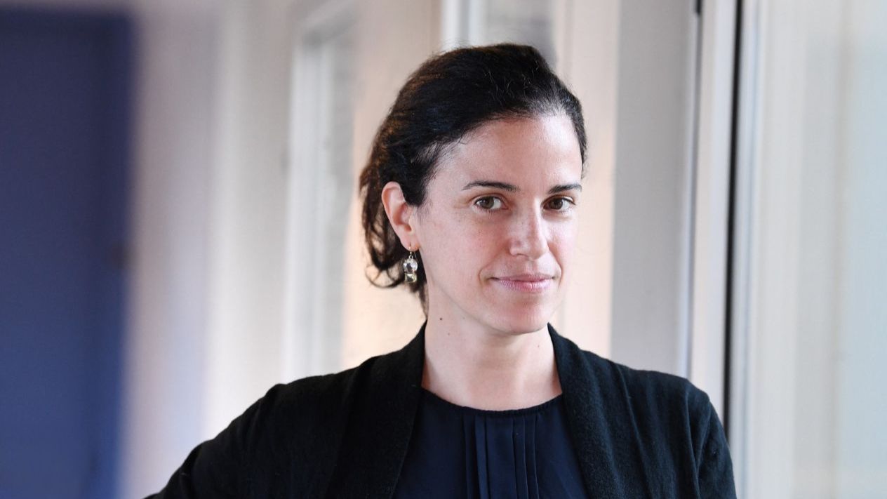 Portrait photo of Professor Elisa Konofagou, a researcher, dressed in a black jacket over a dark blue shirt, looking into the camera in a well-lit hallway with white walls and a blue door.
