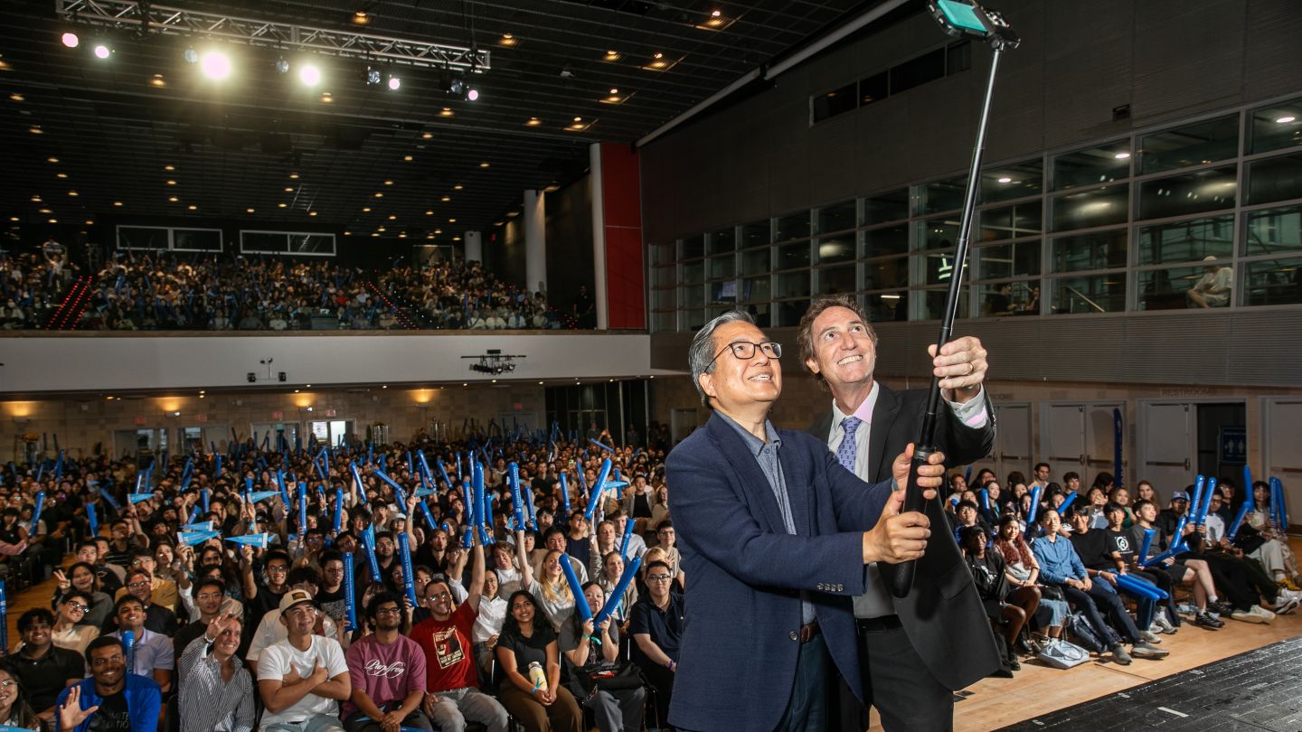 Dean Shih-Fu Chang holds a selfie stick with another man as a large auditorium audience gathers behind them.