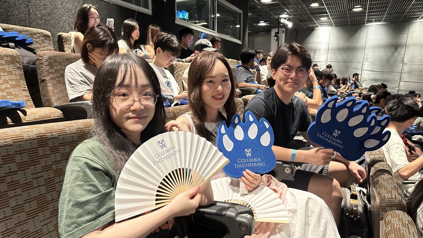 Three seated students hold Columbia Engineering fans and blue foam paw signs in an auditorium.
