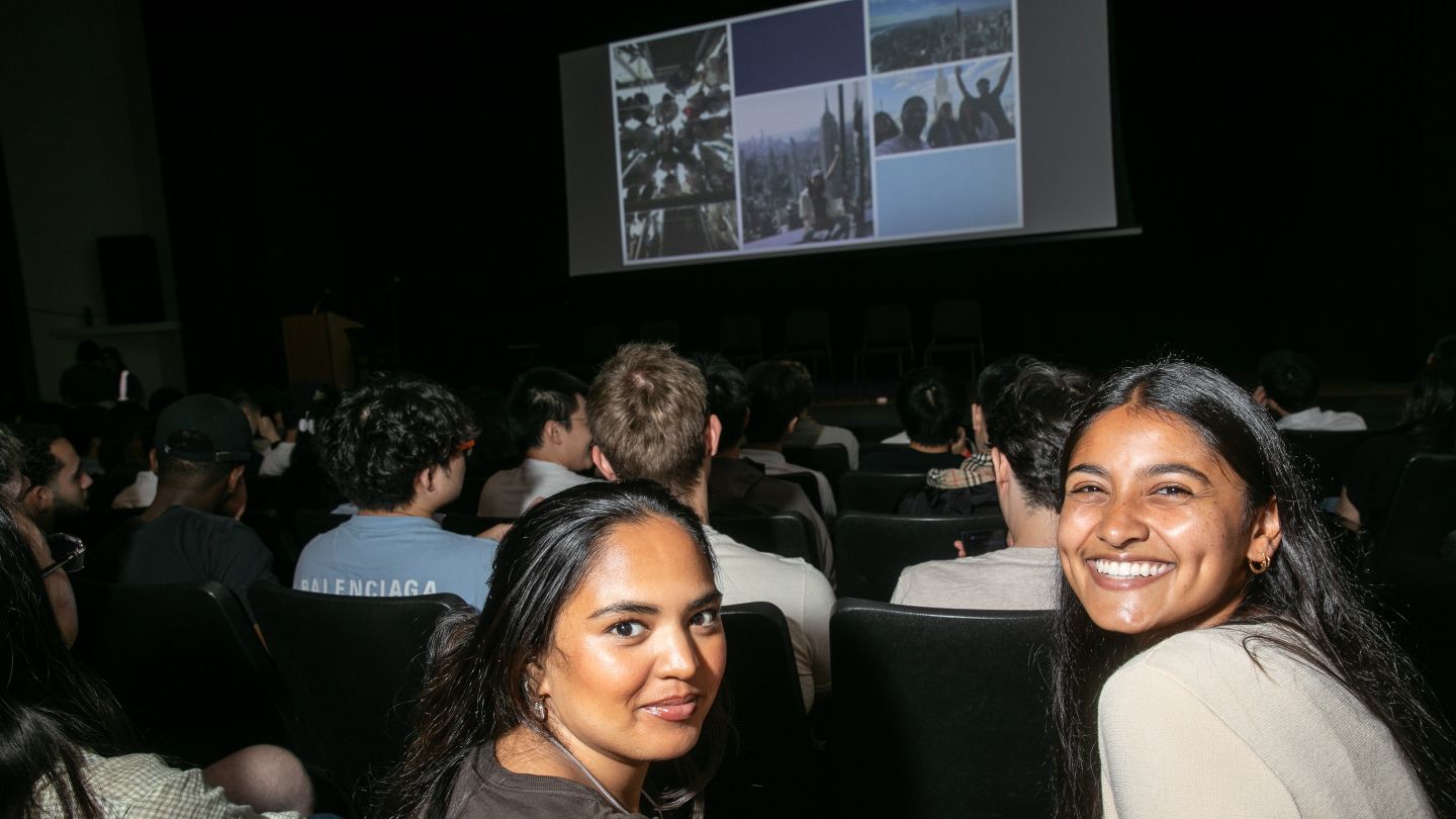 Two students turn toward the camera from their seats as other attendees face a presentation screen.