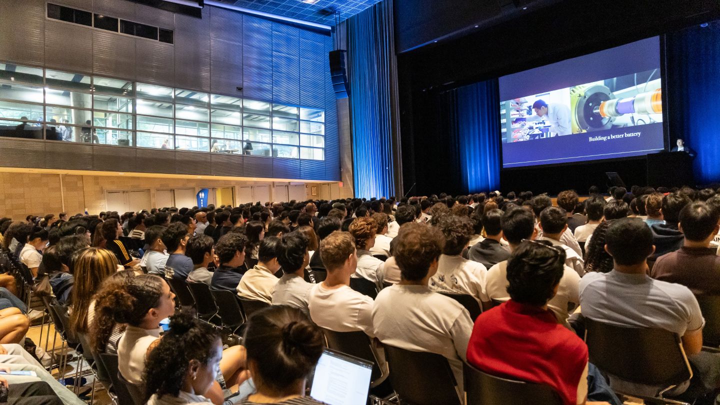 Rows of attendees face a large auditorium screen displaying images of engineering research.