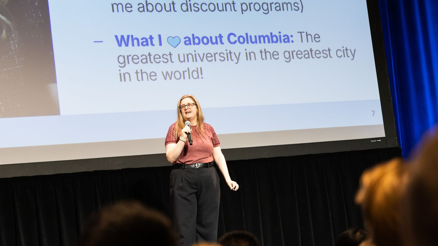 Elizabeth Strauss holds a microphone onstage in front of a projected presentation slide.
