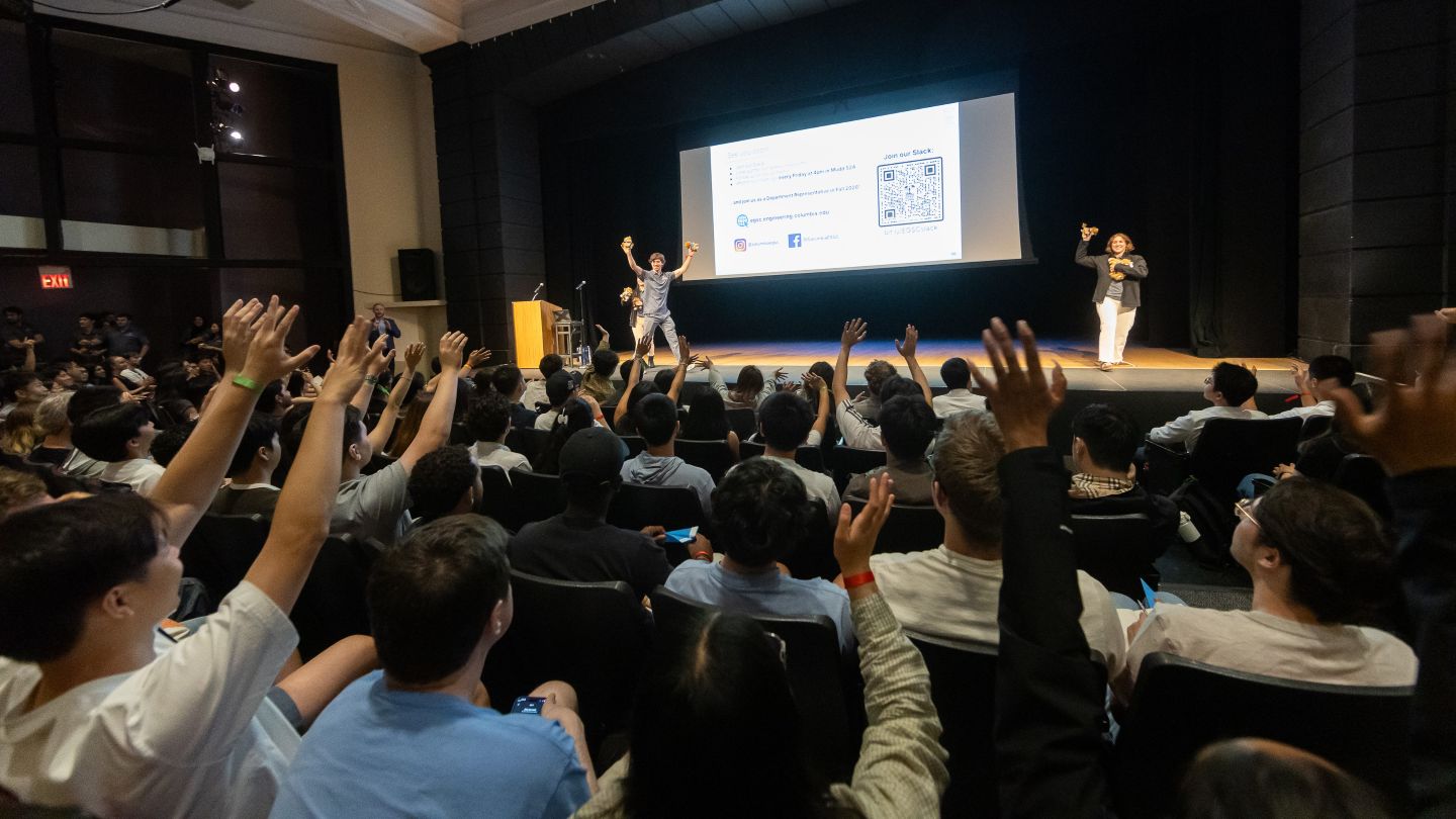 Attendees raise their hands toward two presenters holding small stuffed lions onstage.
