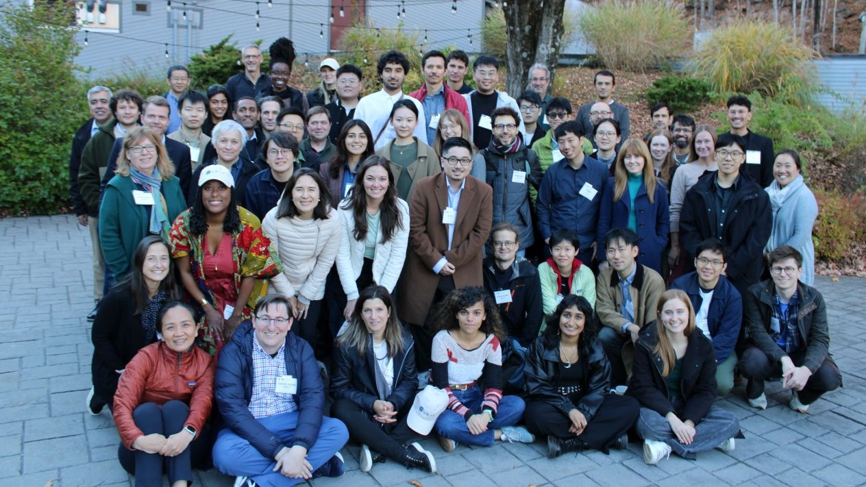 Large group of attendees, some wearing name badges, posed in rows outdoors in front of a building with autumn foliage in the background.