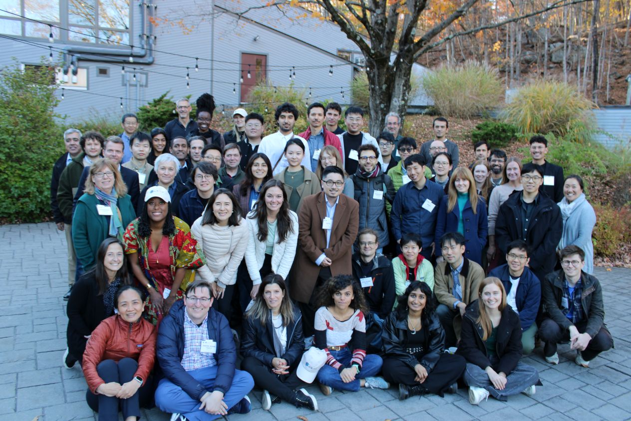 Large group of attendees, some wearing name badges, posed in rows outdoors in front of a building with autumn foliage in the background.