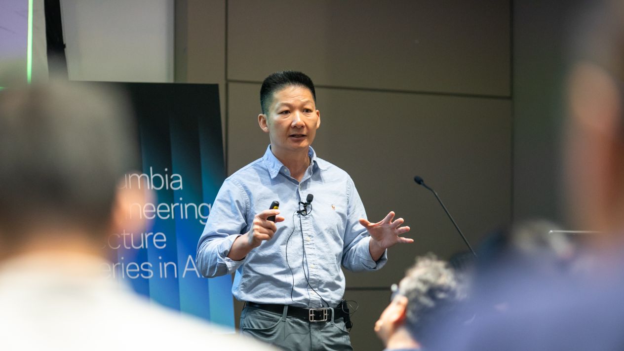Jun Rao speaking and gesturing to an audience during a presentation.