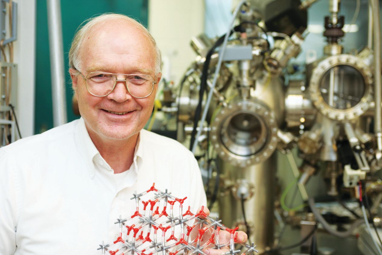 Richard M. Osgood holding a red and silver molecular model in front of laboratory equipment.