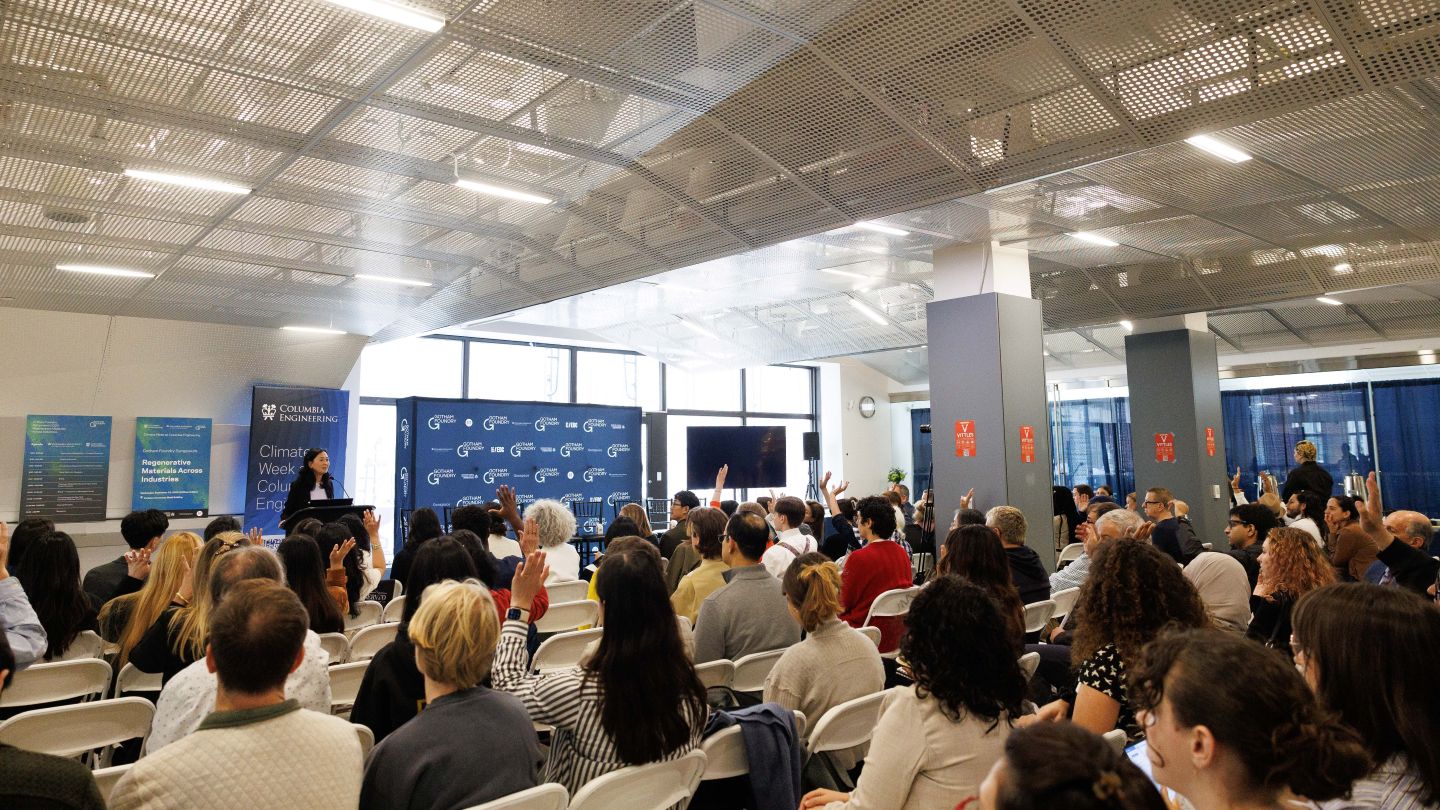 Helen Lu speaking from a podium as audience members raise their hands at the Gotham Foundry symposium.