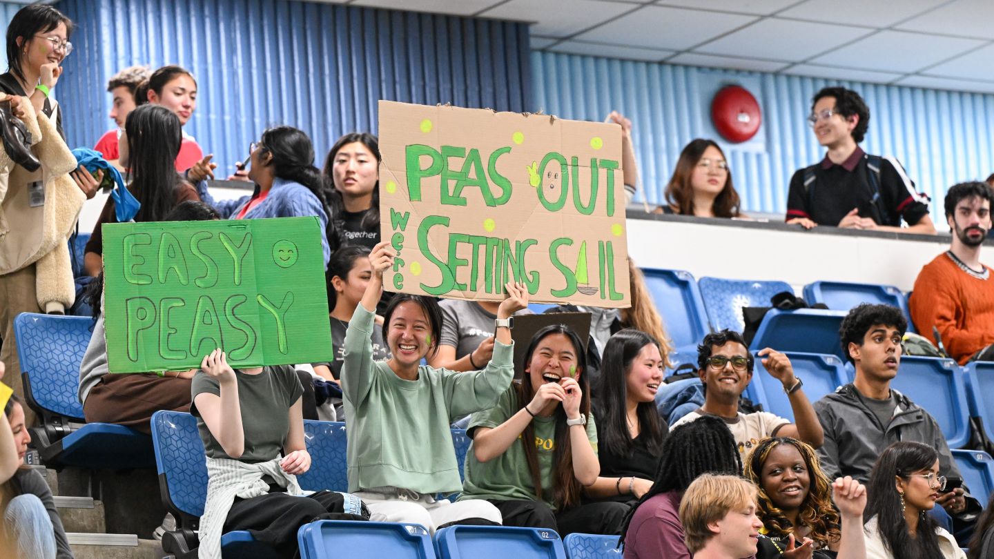 Students cheer from the stands while holding handmade signs during the cardboard boat race at Uris Pool.