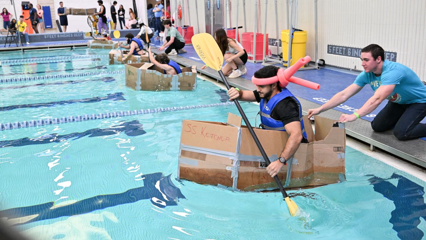 Student wearing a pink balloon hat paddles a cardboard boat as another student pushes it into Uris Pool.