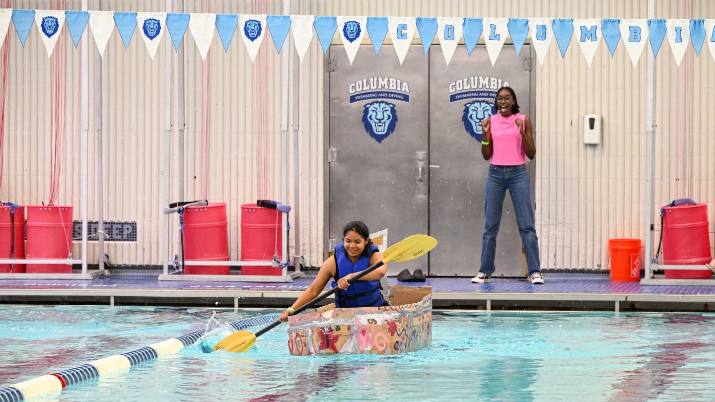 Student paddles a decorated cardboard boat across Uris Pool as another student cheers from the pool deck.